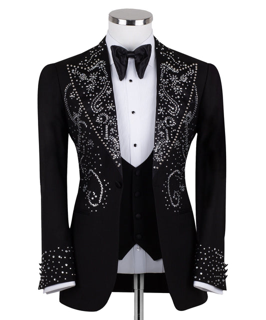 Black embellished three-piece tuxedo with intricate crystal and bead patterns on satin peak lapels, chest, and cuffs. Features a matching vest. Handcrafted luxury suit from Charly Luxury Istanbul.