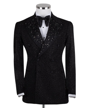Close-up of a black double-breasted tuxedo jacket on a mannequin, showcasing intricate hand-embellishments with beads and crystals on the lapels and body. Features a peak lapel and black buttons, paired with a white shirt and black bow tie. Front view showing the full jacket and details.