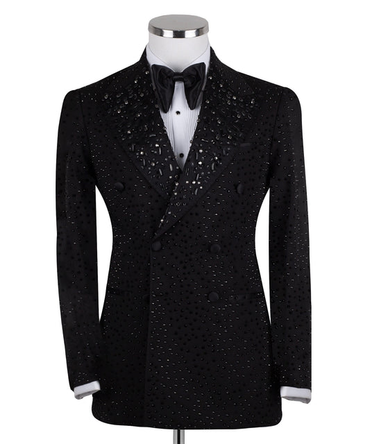 Close-up of a black double-breasted tuxedo jacket on a mannequin, showcasing intricate hand-embellishments with beads and crystals on the lapels and body. Features a peak lapel and black buttons, paired with a white shirt and black bow tie. Front view showing the full jacket and details.