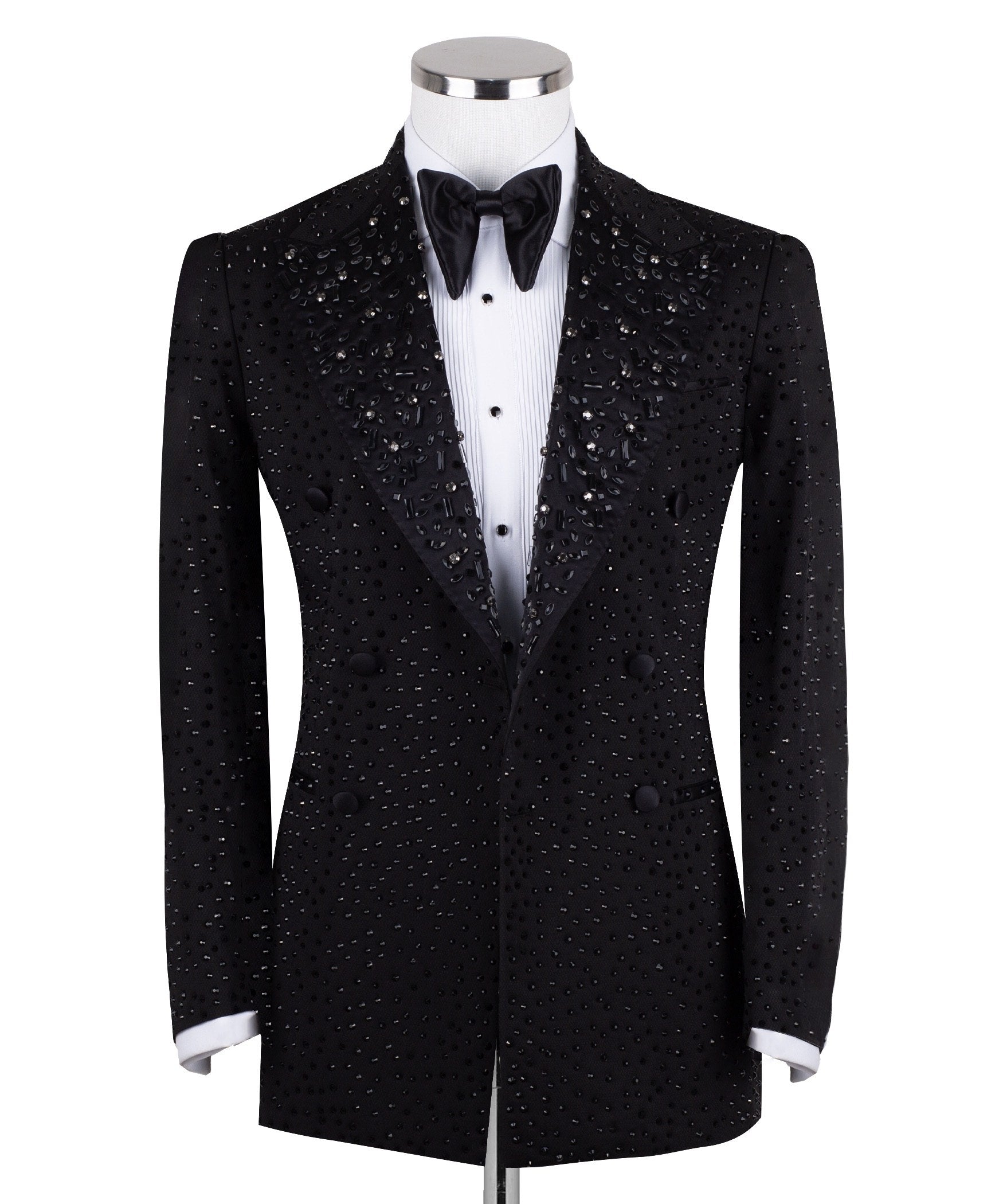 Close-up of a black double-breasted tuxedo jacket on a mannequin, showcasing intricate hand-embellishments with beads and crystals on the lapels and body. Features a peak lapel and black buttons, paired with a white shirt and black bow tie. Front view showing the full jacket and details.
