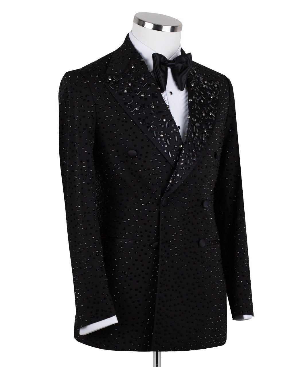 Close-up of a black double-breasted tuxedo jacket on a mannequin, showcasing intricate hand-embellishments with beads and crystals on the lapels and body. Features a peak lapel and black buttons, paired with a white shirt and black bow tie. Front view showing the full jacket and details.