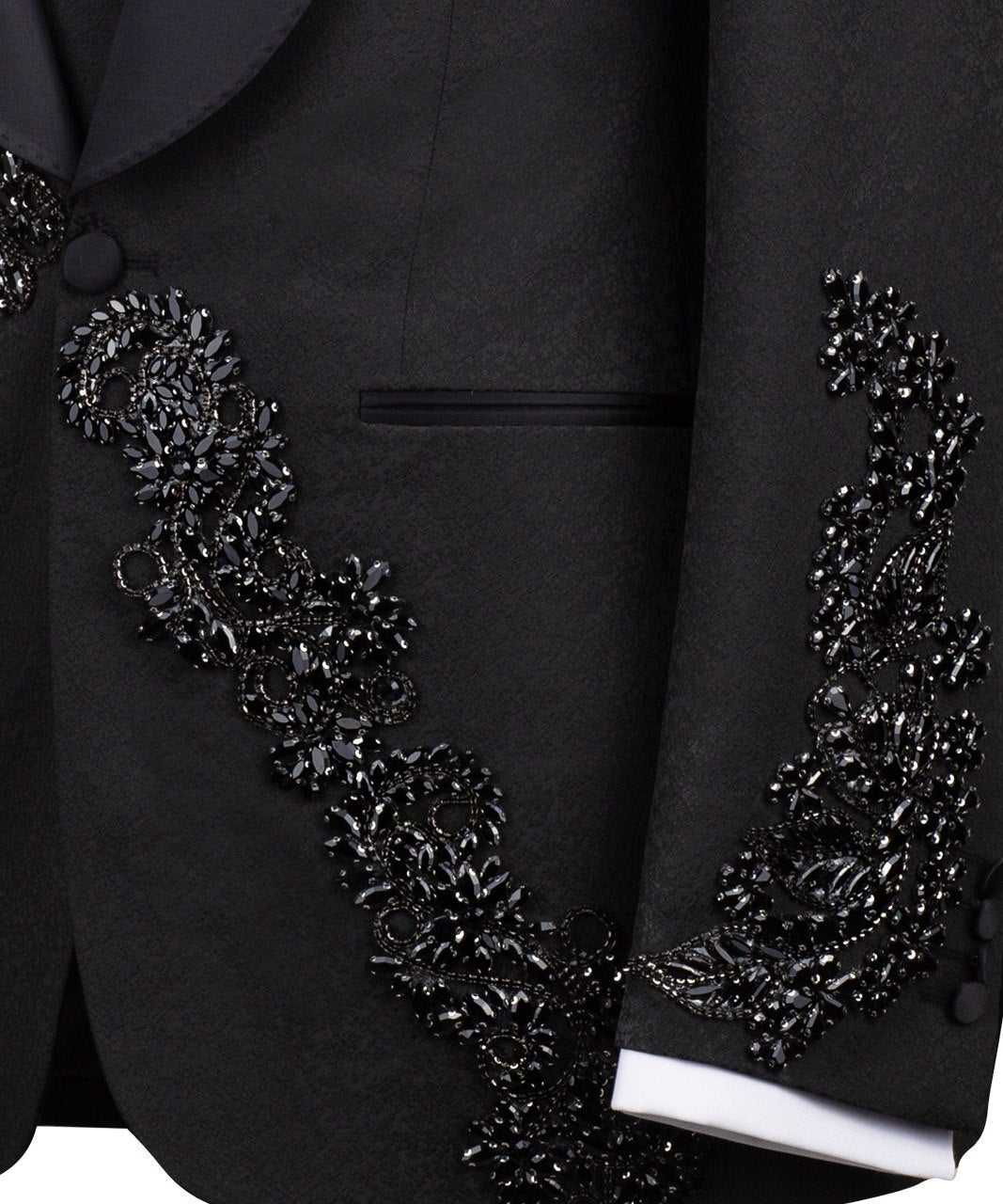 Black slim fit hand-embellished tuxedo on a mannequin, featuring a satin peak lapel, matching vest, and intricate black beadwork detailing on the shoulder, lapel, and sleeves. The tuxedo fabric has a subtle jacquard texture, showcasing master tailoring from Istanbul.
