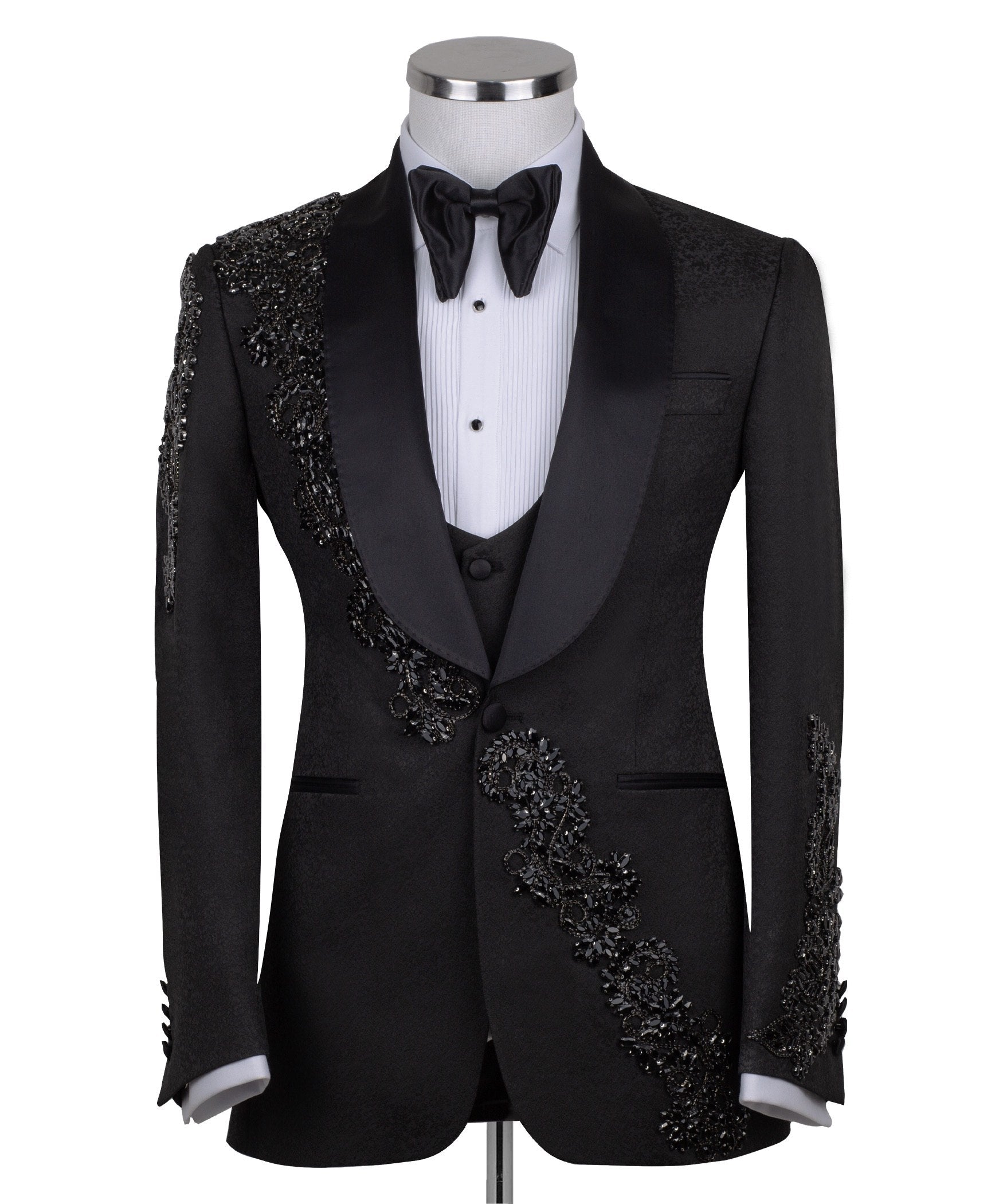 Black slim fit hand-embellished tuxedo on a mannequin, featuring a satin peak lapel, matching vest, and intricate black beadwork detailing on the shoulder, lapel, and sleeves. The tuxedo fabric has a subtle jacquard texture, showcasing master tailoring from Istanbul.