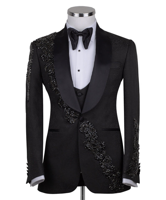 Black slim fit hand-embellished tuxedo on a mannequin, featuring a satin peak lapel, matching vest, and intricate black beadwork detailing on the shoulder, lapel, and sleeves. The tuxedo fabric has a subtle jacquard texture, showcasing master tailoring from Istanbul.