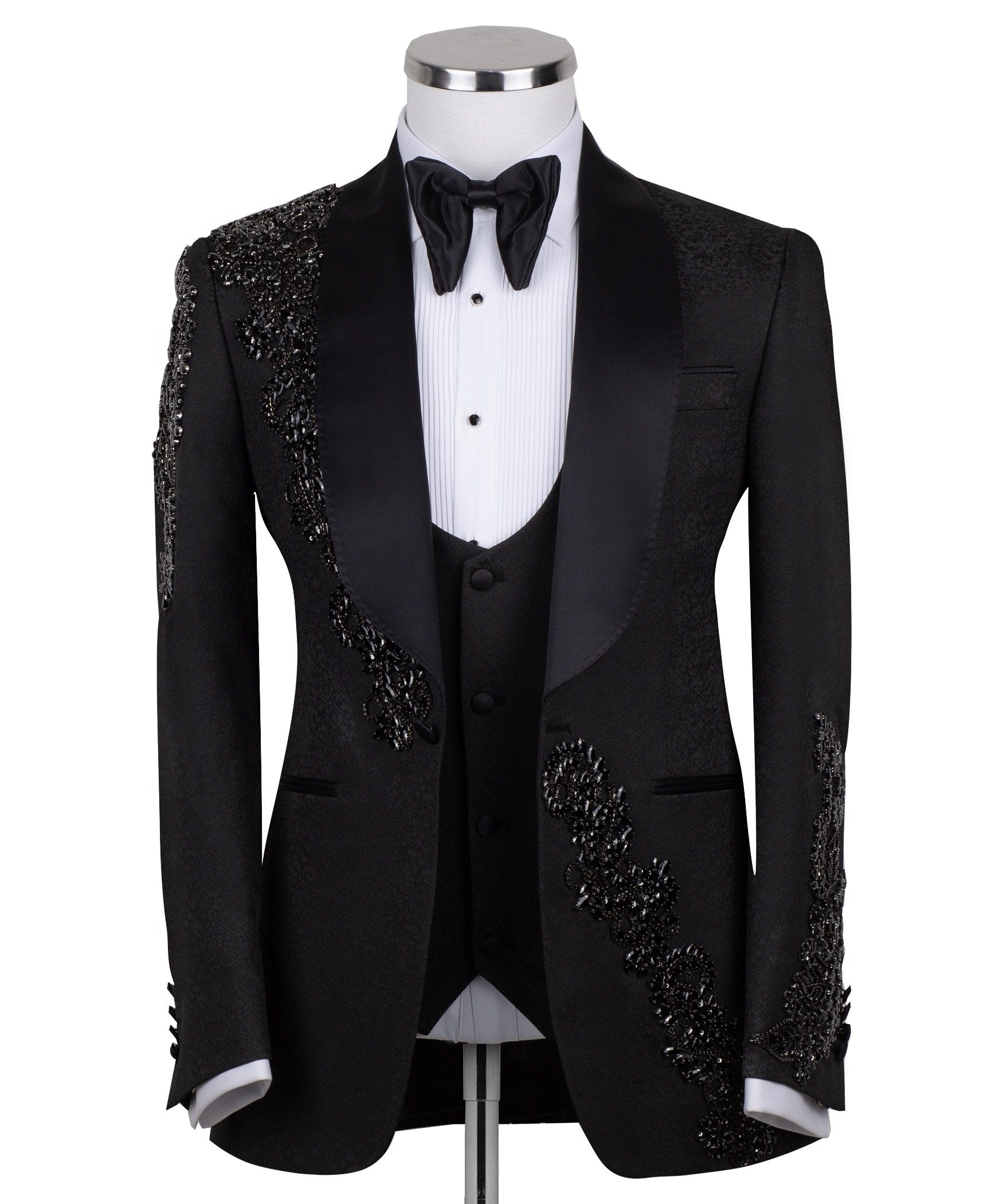 Black slim fit hand-embellished tuxedo on a mannequin, featuring a satin peak lapel, matching vest, and intricate black beadwork detailing on the shoulder, lapel, and sleeves. The tuxedo fabric has a subtle jacquard texture, showcasing master tailoring from Istanbul.