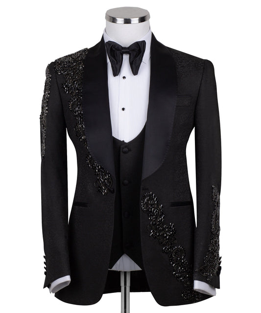 Black slim fit hand-embellished tuxedo on a mannequin, featuring a satin peak lapel, matching vest, and intricate black beadwork detailing on the shoulder, lapel, and sleeves. The tuxedo fabric has a subtle jacquard texture, showcasing master tailoring from Istanbul.
