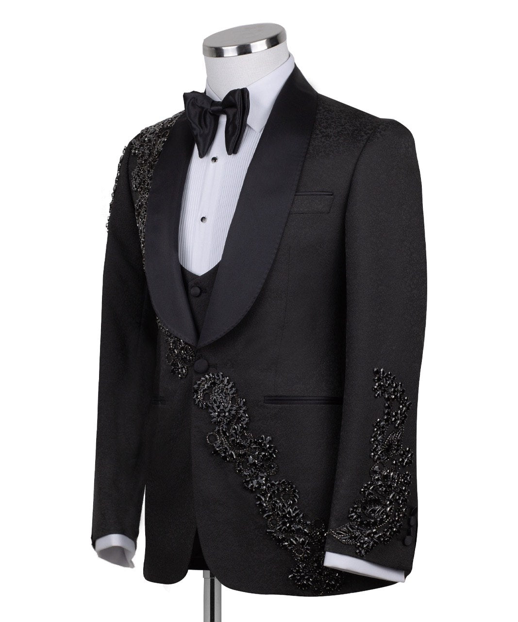 Black slim fit hand-embellished tuxedo on a mannequin, featuring a satin peak lapel, matching vest, and intricate black beadwork detailing on the shoulder, lapel, and sleeves. The tuxedo fabric has a subtle jacquard texture, showcasing master tailoring from Istanbul.