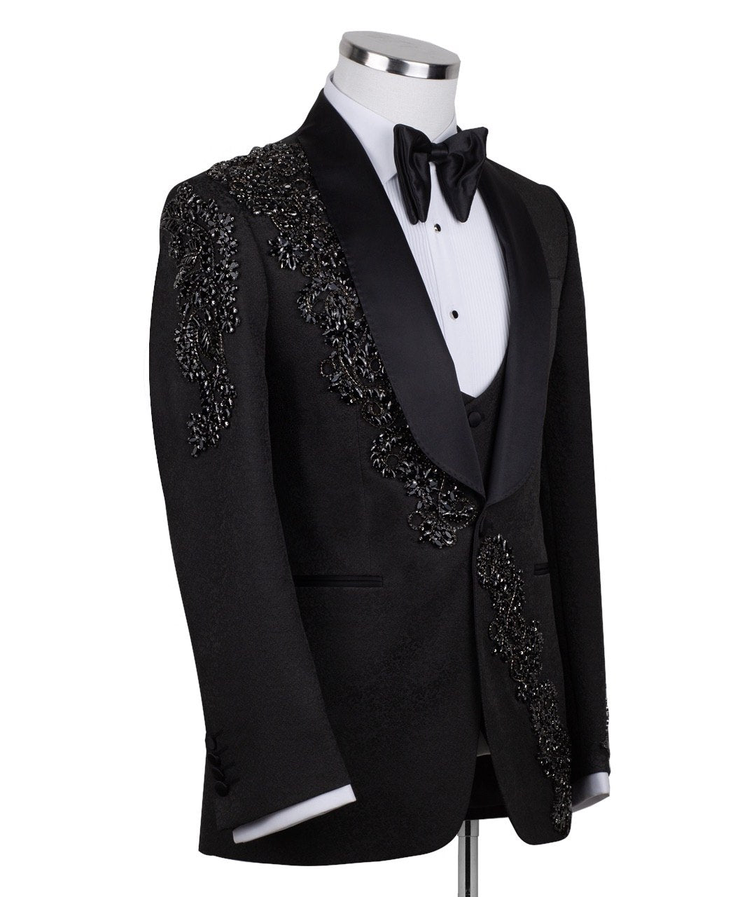 Black slim fit hand-embellished tuxedo on a mannequin, featuring a satin peak lapel, matching vest, and intricate black beadwork detailing on the shoulder, lapel, and sleeves. The tuxedo fabric has a subtle jacquard texture, showcasing master tailoring from Istanbul.