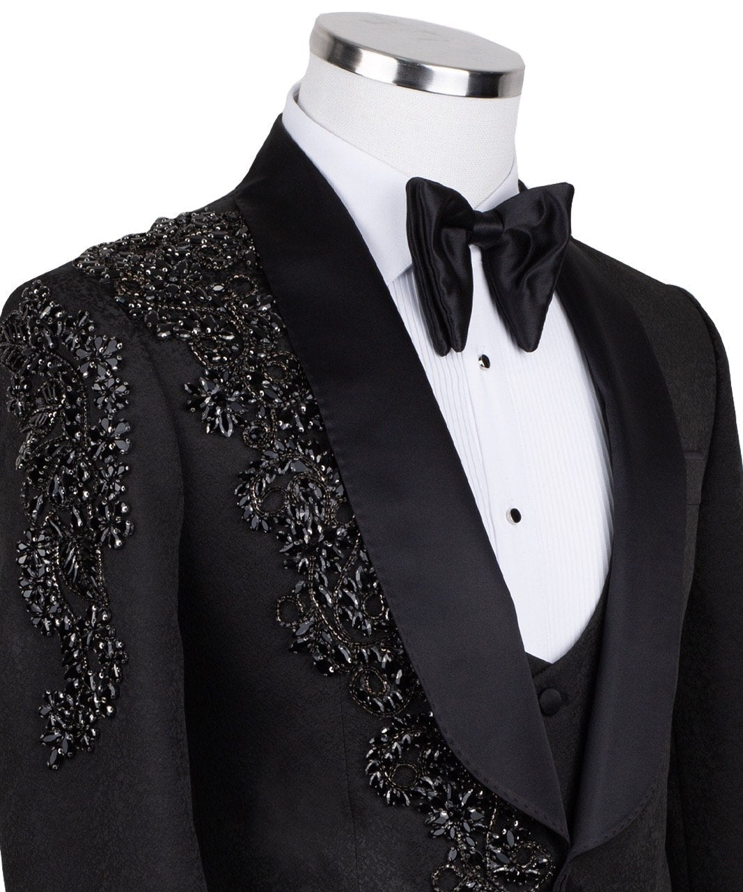 Black slim fit hand-embellished tuxedo on a mannequin, featuring a satin peak lapel, matching vest, and intricate black beadwork detailing on the shoulder, lapel, and sleeves. The tuxedo fabric has a subtle jacquard texture, showcasing master tailoring from Istanbul.