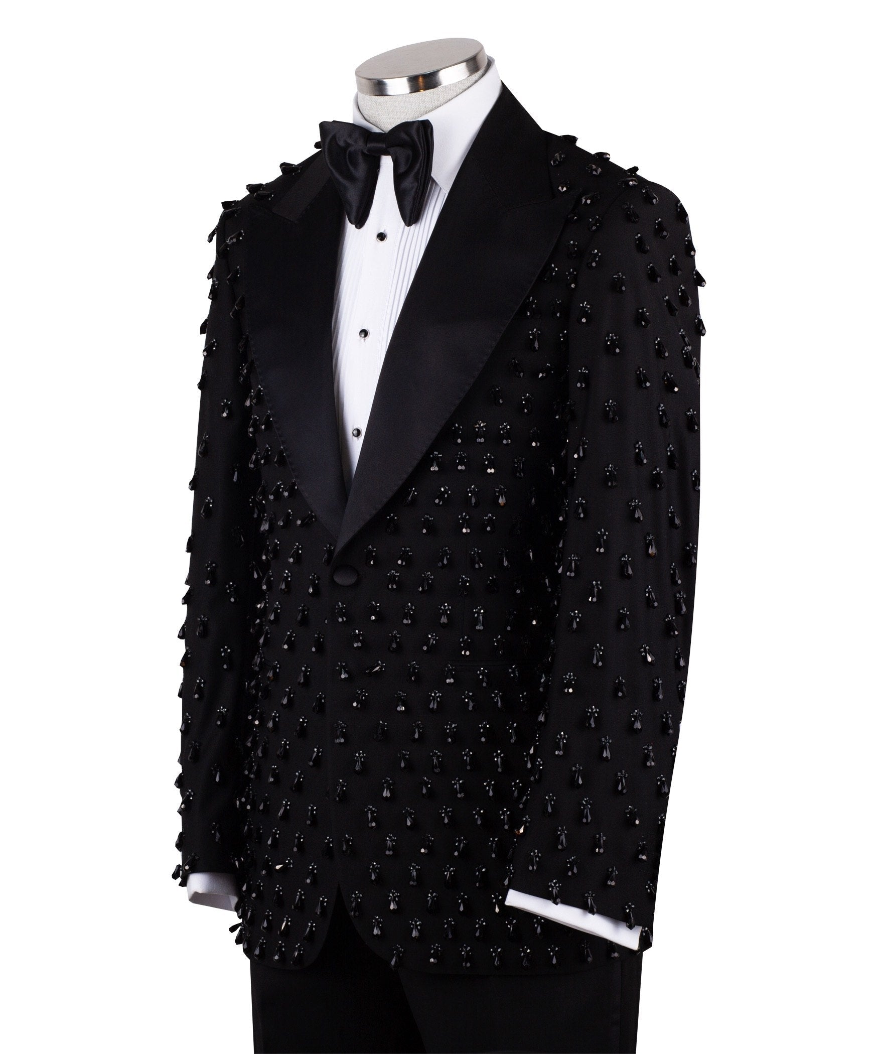 Front view of a black hand-embellished luxury tuxedo jacket with satin peak lapels and matching trousers on a mannequin. Side view close-up of the jacket's intricate black bead embellishments. Close-up of the tuxedo trousers with a satin side stripe and high-waisted detail.