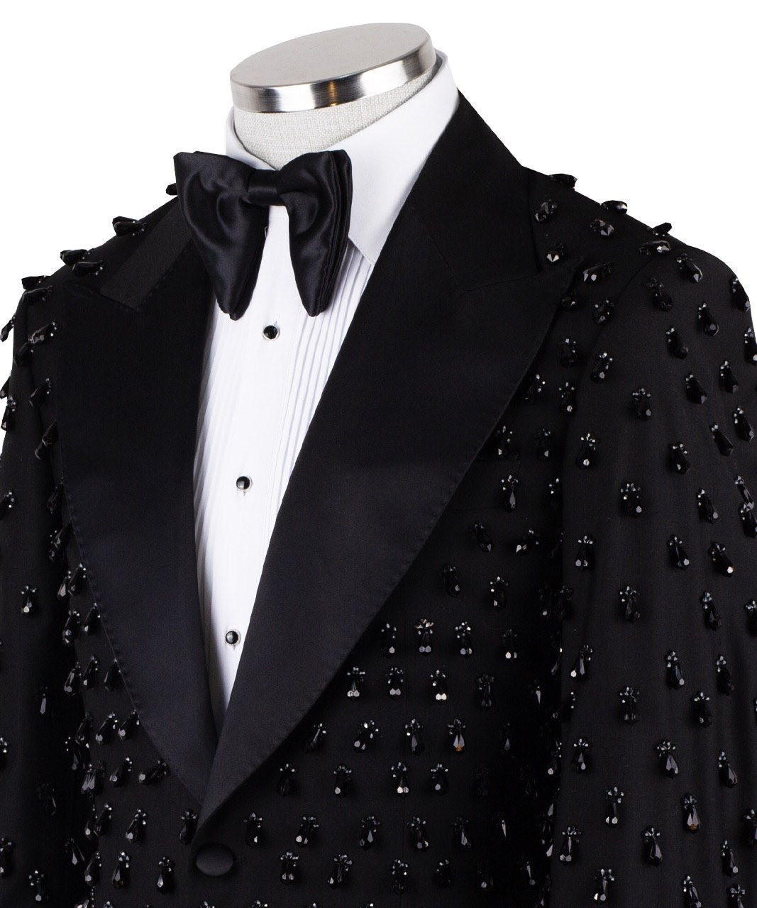 Front view of a black hand-embellished luxury tuxedo jacket with satin peak lapels and matching trousers on a mannequin. Side view close-up of the jacket's intricate black bead embellishments. Close-up of the tuxedo trousers with a satin side stripe and high-waisted detail.