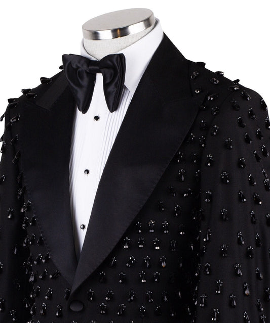 Front view of a black hand-embellished luxury tuxedo jacket with satin peak lapels and matching trousers on a mannequin. Side view close-up of the jacket's intricate black bead embellishments. Close-up of the tuxedo trousers with a satin side stripe and high-waisted detail.