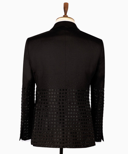 Front view of a black double-breasted tuxedo jacket featuring intricate hand-embellished studs on the peak lapels, shoulders, sleeves, and lower body, styled with matching black trousers with a satin side stripe. Handcrafted luxury from Charly Luxury Istanbul.