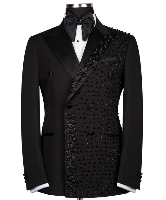 Front view of a black double-breasted tuxedo jacket with satin peak lapels and intricate hand-embellished black beading on the right side. Paired with matching trousers featuring a satin side stripe. High-end bespoke formal wear, handcrafted in Istanbul.