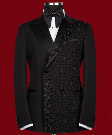 Front view of a black double-breasted tuxedo jacket with satin peak lapels and intricate hand-embellished black beading on the right side. Paired with matching trousers featuring a satin side stripe. High-end bespoke formal wear, handcrafted in Istanbul.