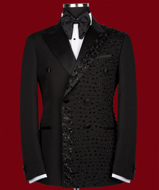 Front view of a black double-breasted tuxedo jacket with satin peak lapels and intricate hand-embellished black beading on the right side. Paired with matching trousers featuring a satin side stripe. High-end bespoke formal wear, handcrafted in Istanbul.