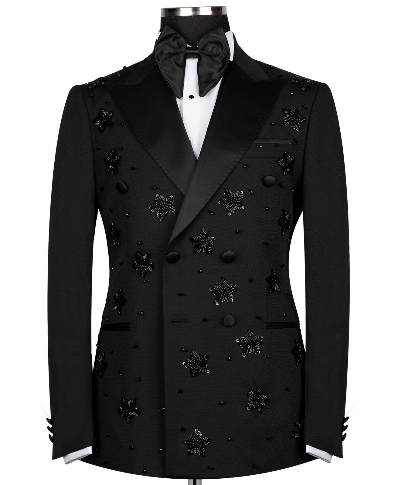 Front view of a black double-breasted tuxedo with satin peak lapels, adorned with intricate black beaded starburst embellishments, worn by a mannequin. The matching trousers with satin side stripe are also shown. This luxurious evening wear is handcrafted in Istanbul by Charly Luxury.