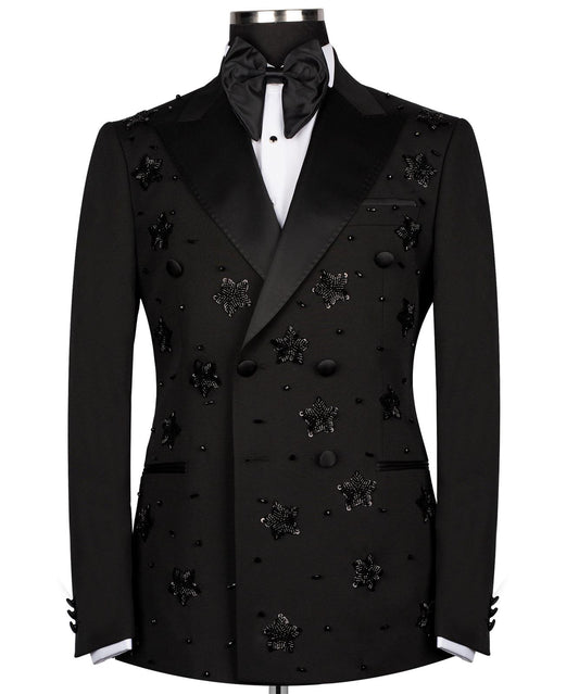 Front view of a black double-breasted tuxedo with satin peak lapels, adorned with intricate black beaded starburst embellishments, worn by a mannequin. The matching trousers with satin side stripe are also shown. This luxurious evening wear is handcrafted in Istanbul by Charly Luxury.