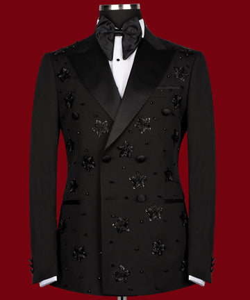 Front view of a black double-breasted tuxedo with satin peak lapels, adorned with intricate black beaded starburst embellishments, worn by a mannequin. The matching trousers with satin side stripe are also shown. This luxurious evening wear is handcrafted in Istanbul by Charly Luxury.
