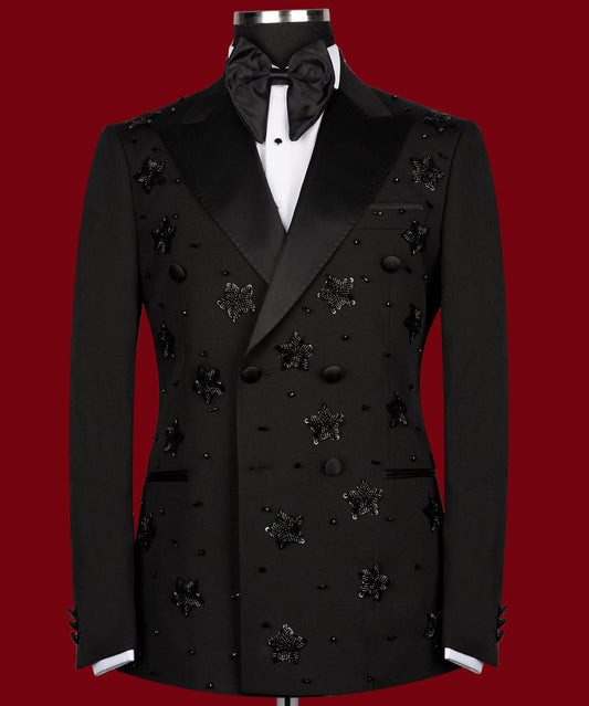 Front view of a black double-breasted tuxedo with satin peak lapels, adorned with intricate black beaded starburst embellishments, worn by a mannequin. The matching trousers with satin side stripe are also shown. This luxurious evening wear is handcrafted in Istanbul by Charly Luxury.